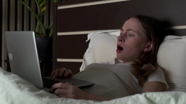 La mujer se acuesta en la cama con una computadora portátil y bosteza varias veces, luego se duerme. Intento trabajar de noche.. — Vídeo de stock