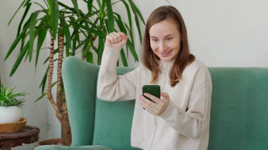 Kanepede oturan, akıllı telefon kullanan bir kadın. Heyecanlı kadın ekrana bak, telefon oyununda kazanıyor.