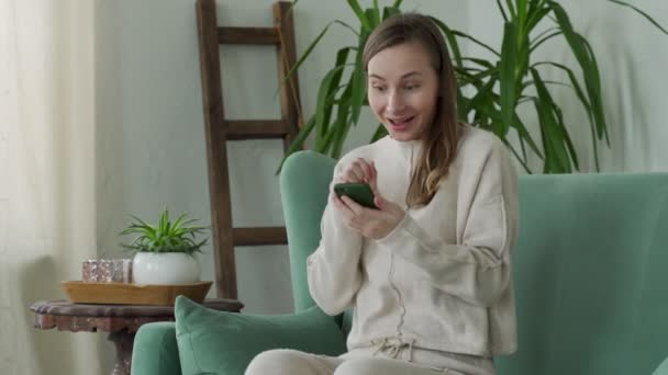 Mujer joven usando el teléfono, celebrando la victoria de la lotería en línea, leyendo buen mensaje, sentado en el sofá en casa — Vídeos de Stock