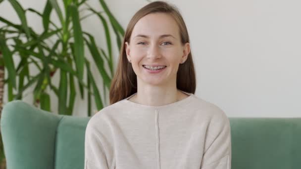 Een gelukkige jonge vrouw met een beugel glimlacht naar de camera terwijl ze op een stoel zit — Stockvideo