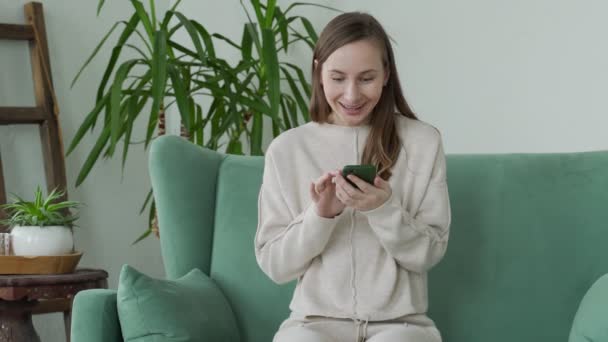 Junge Frau telefoniert, feiert Online-Lottogewinn, liest gute Nachricht, sitzt zu Hause auf dem Sofa — Stockvideo