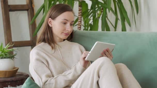 Mooie jonge brunette vrouw met behulp van tablet zitten op de bank thuis in de woonkamer — Stockvideo