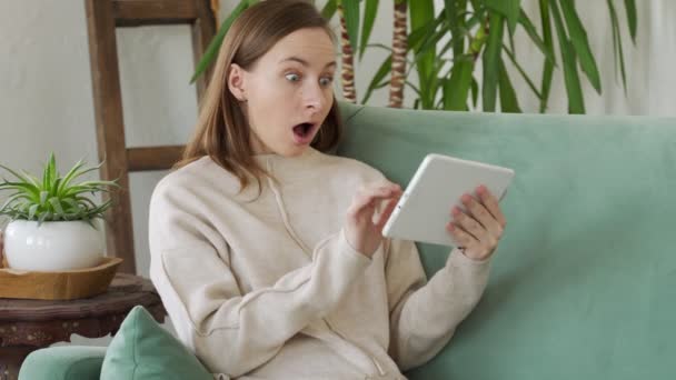 Attractive woman sitting at home on sofa with her tablet computer, celebrating success — Stock Video