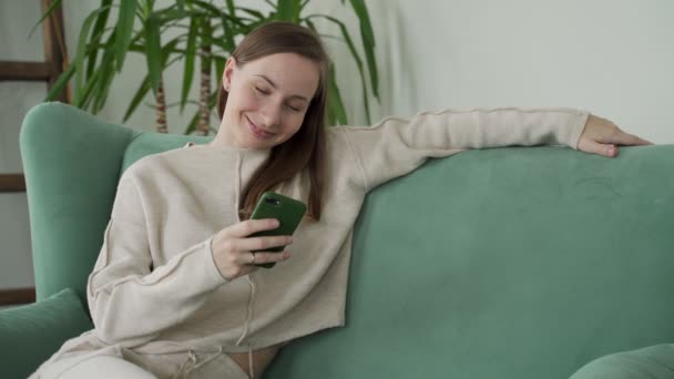 Donna sorridente si rilassa seduto sul divano a casa utilizzando i messaggi di testo del telefono cellulare per chattare con un amico — Video Stock