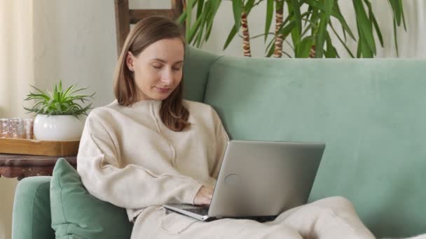 Молода брюнетка сидить на дивані і робить проект на ноутбуці в затишній вітальні вдома — стокове відео