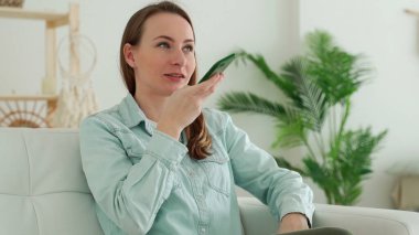 Evdeki genç esmer kadın kanepede oturup telefonla sesli mesaj yolluyor.