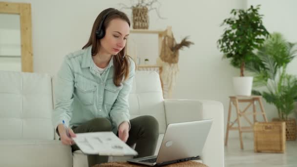 Mulher vestindo fone de ouvido com microfone, usando laptop em casa, sentado no sofá na sala de estar — Vídeo de Stock