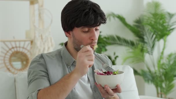 Mladý bruneta muž jíst čerstvý zeleninový salát, zatímco sedí na gauči doma — Stock video