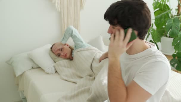 Zamartwiony mąż dzwoni do lekarza z chorą żoną leżącą w tle. Mąż konsultant lekarza na telefon — Wideo stockowe
