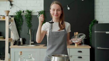 Mutlu kadın bir kepçeyle çorbanın tadına bakar ve mutfakta gülümser.