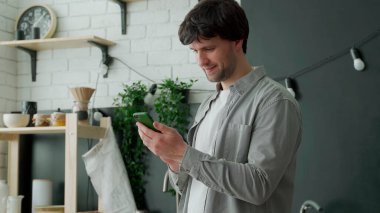 Adam mutfakta telefonla internette sörf yapıyor. Mutfakta duran ve akıllı telefon kullanan bir adamın portresi.