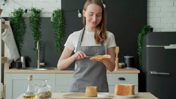 Uma mulher está em um avental na cozinha e espalha manteiga de amendoim em torradas — Fotografia de Stock