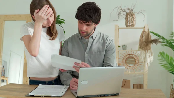 Stressed couple looking frustrated, having no money to pay off their debts, sitting at table with papers, calculator and laptop computer and reading document from bank