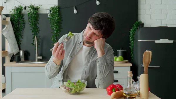 Unhappy man eating vegetable salad at table in kitchen. Displeased young man eating green leaf lettuce — Stock Photo, Image