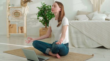 Genç kadın evde yerde otururken dizüstü bilgisayar kullanarak yoga egzersizleri yapıyor.