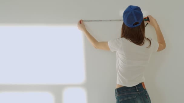 Mujer joven con una gorra azul con cinta métrica dibuja etiquetas en una pared blanca en su apartamento — Vídeos de Stock