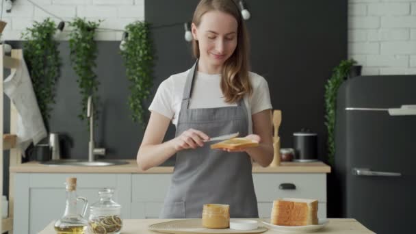 Una donna sta in un grembiule in cucina e spalma burro di arachidi sul pane tostato — Video Stock