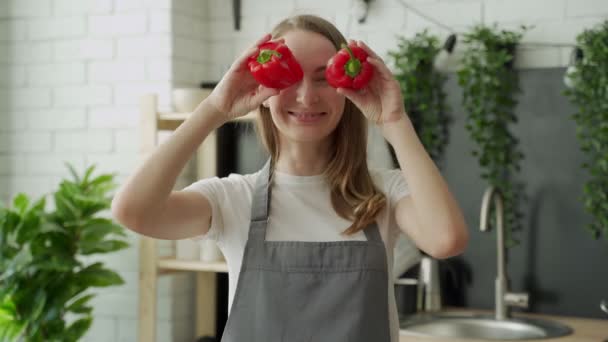 Heureuse jeune femme sourit et s'amuse à jouer avec le poivron rouge devant ses yeux dans la cuisine de la maison — Video