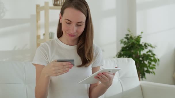 Mujer sonriente en el sofá usando una tableta y tarjeta bancaria hace compras en línea en casa en la sala de estar — Vídeos de Stock