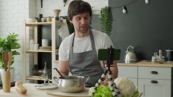 Jovem chef em um avental filmando-se para um blog de culinária enquanto cozinha uma refeição saudável com legumes na cozinha — Vídeo de Stock