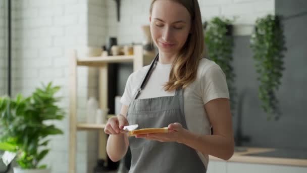 En kvinna står i ett förkläde i köket och sprider jordnötssmör på rostat bröd — Stockvideo