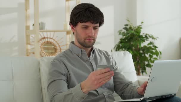El joven está sentado en el sofá de la sala de estar, haciendo compras en línea a través de un ordenador portátil, sosteniendo una tarjeta de crédito, pagando las compras en una tienda en línea que descansa en casa — Vídeo de stock