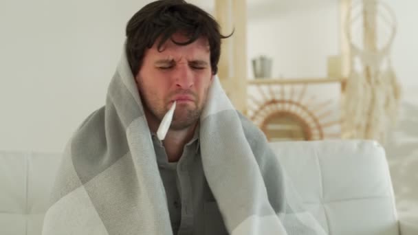Egy megfázott férfi ül a kanapén egy hőmérővel a szájában. Egy ember otthon influenzás, megméri a lázát. — Stock videók