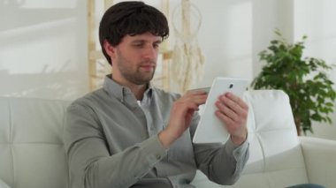 Tablet bilgisayarı ve evde kredi kartı olan gülümseyen siyah saçlı bir adam kanepede otururken internetten alışveriş yapıyor.