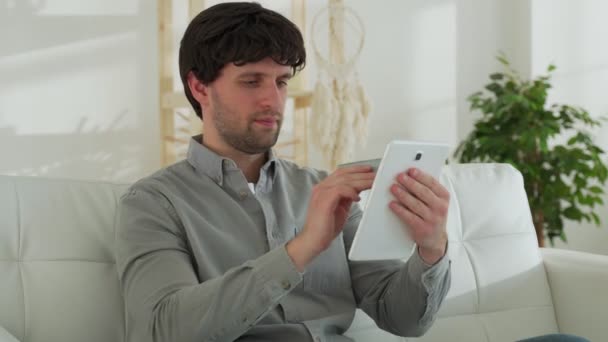 Ένας χαμογελαστός μελαχρινός άντρας με έναν υπολογιστή tablet και μια πιστωτική κάρτα στο σπίτι κάνει online αγορές ενώ κάθεται στον καναπέ — Αρχείο Βίντεο