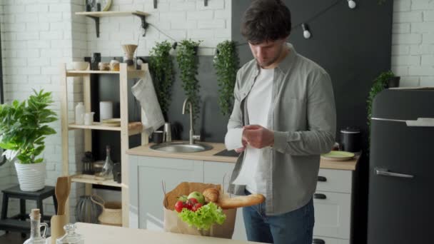 Ein junger Mann mit einer Tüte Lebensmittel kontrolliert und prüft einen Warenbeleg, nachdem er in einem Lebensmittelgeschäft Lebensmittel gekauft hat. Mann studiert Lebensmittelkontrolle, während sie in ihrer Küche steht — Stockvideo