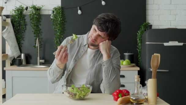 Hombre infeliz comiendo ensalada de verduras en la mesa en la cocina. Joven disgustado comiendo lechuga de hoja verde — Vídeos de Stock