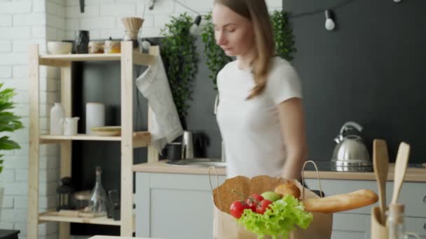 Mujer se para en la cocina cerca de una bolsa de papel llena de comida fresca y utiliza una aplicación para teléfonos inteligentes para entregarlo al supermercado — Vídeo de stock