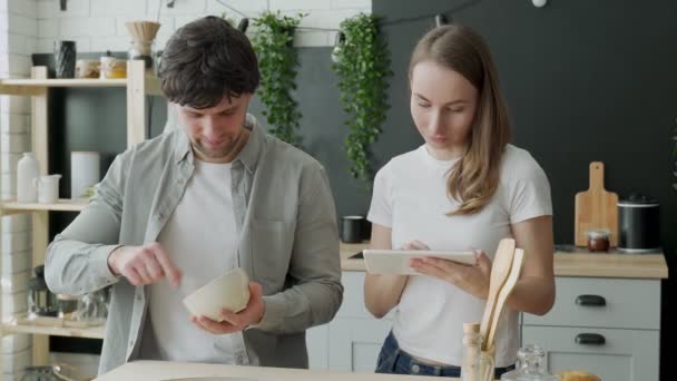 Молодая супружеская пара использует цифровой планшет и улыбается во время приготовления пищи на кухне дома — стоковое видео