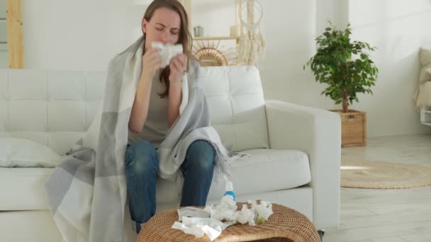 Una mujer envuelta en una manta se siente enferma con un resfriado y fiebre en casa, enferma de gripe, sentada en el sofá — Vídeo de stock