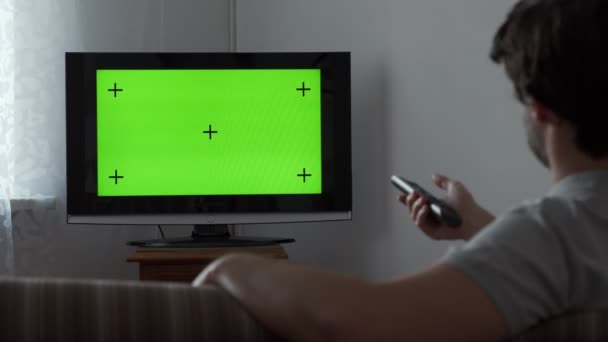 Junger Mann sitzt auf einem Sofa und sieht fern mit grünem Bildschirm. — Stockvideo