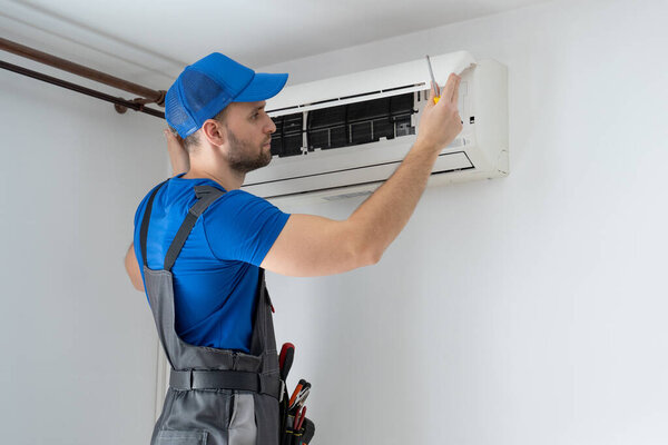Male technician in overalls and a blue cap repairs an air conditioner on the wall