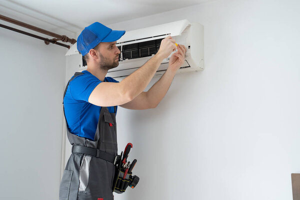 Male technician in overalls and a blue cap repairs an air conditioner on the wall