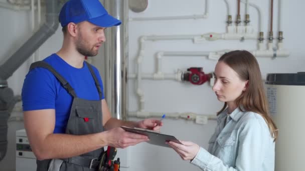 A female client signs a bill from a male plumber standing in the boiler room — Stock Video