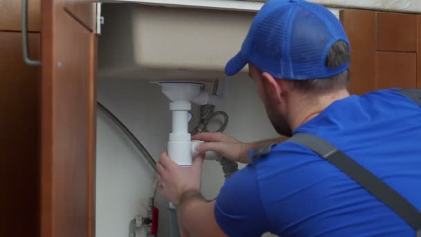 Jovem reparador com ferramentas em suas mãos e um boné azul está consertando a pia na cozinha — Vídeo de Stock