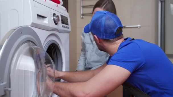 Reparatören reparerar en tvättmaskin framför en kvinna. En man kommunicerar med ägaren av en tvättmaskin — Stockvideo