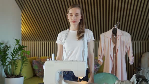 若い女性のドレスメーカーは、ワークショップでポーズ、腕はミシンの背景に交差 — ストック動画