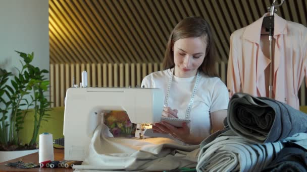 Giovane cucitrice prende appunti sulle misure di abbigliamento su un taccuino in un laboratorio di cucito. — Video Stock