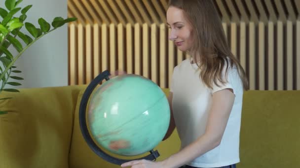 Jovem mulher enquanto se senta no sofá em casa segurando um globo mundial escolher uma rota de viagem. — Vídeo de Stock