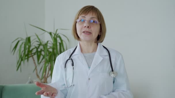 Ärztin in weißer Arztuniform mit Stethoskop spricht Videokonferenz mit Patientin, schaut in die Kamera. — Stockvideo