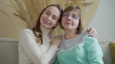 Sevgiyle annesini kucaklayan, mutlu yüz ifadeleri takınan, kameraya geniş bir gülümsemeyle gülümseyen şık hippi gözlüklü sevimli genç kızın yatay görüntüsü.