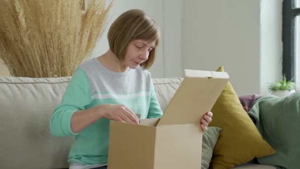 Mulher de meia idade desempacotar caixa de papelão, sentado no sofá em casa. Senior mulher segurar caixa de papelão aberta sentar no sofá na sala de estar — Vídeo de Stock