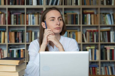 Düşünceli öğrenci kütüphanede oturur ve elinde kalemle sınavları düşünür.