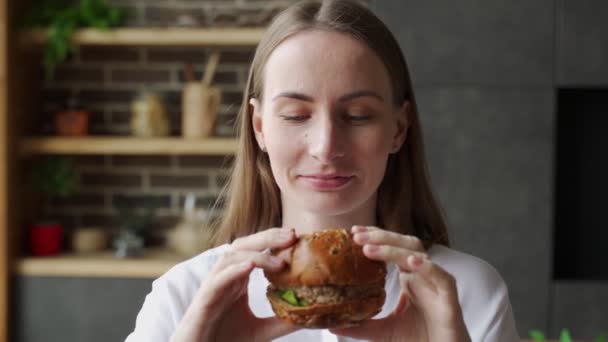 Mujer hambrienta comiendo una hamburguesa sentada en la cocina — Vídeo de stock