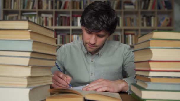 Studente uomo si siede a un tavolo in biblioteca, disseminato di libri. — Video Stock