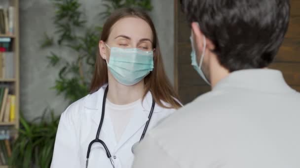 Doktor en kvinna i medicinsk mask pratar med en manlig patient som sitter i en soffa — Stockvideo